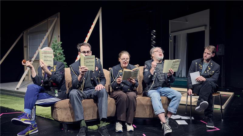 Fünf Personen sitzen auf einem Sofa auf einer Bühne und halten jeweils ein Buch oder Heft in der Hand.