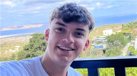 Mann mit lockigem Haar in weißem T-Shirt vor Balkon mit Blick auf bewaldete Landschaft und Wasser im Hintergrund