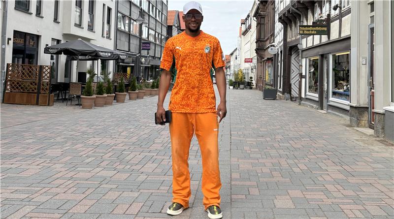 Ein Mann steht auf einer gepflasterten Fußgängerzone in orangefarbener Sportkleidung und hält eine schwarze Tasche in der rechten Hand.