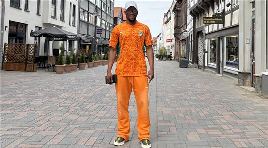Ein Mann steht auf einer gepflasterten Fußgängerzone in orangefarbener Sportkleidung und hält eine schwarze Tasche in der rechten Hand.