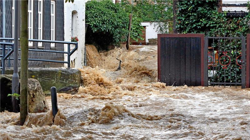 Stark verschmutztes Hochwasser strömt durch einen engen, von Gebäuden und Zäunen gesäumten Bereich.