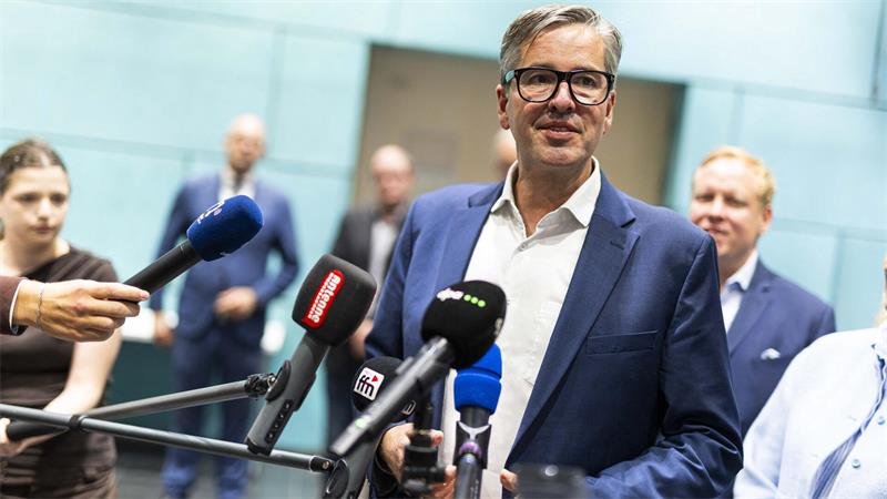 Mann mit Brille und blauem Sakko spricht vor mehreren Mikrofonen, im Hintergrund unscharfe Personen