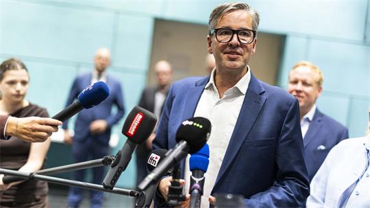 Mann mit Brille und blauem Sakko spricht vor mehreren Mikrofonen, im Hintergrund unscharfe Personen