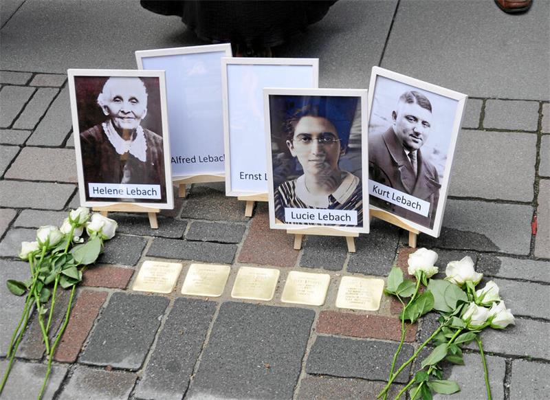 Porträts von vier Personen auf kleinen Staffeleien vor Stolpersteinen auf einem Gehweg, umgeben von weißen Rosen