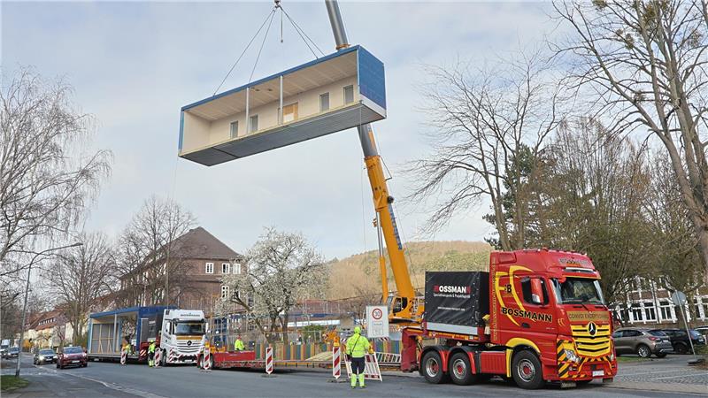 Kran hebt großen rechteckigen Container über Straße mit mehreren Lastwagen und Bauarbeitern in Schutzkleidung