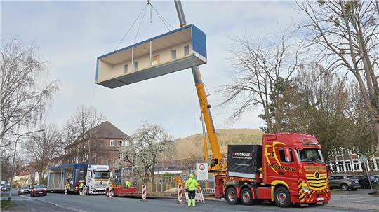 Kran hebt großen rechteckigen Container über Straße mit mehreren Lastwagen und Bauarbeitern in Schutzkleidung