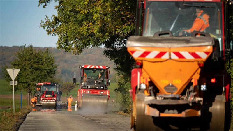 Straßenbauarbeiten mit mehreren Baumaschinen und Arbeitern auf einer Landstraße unter Bäumen