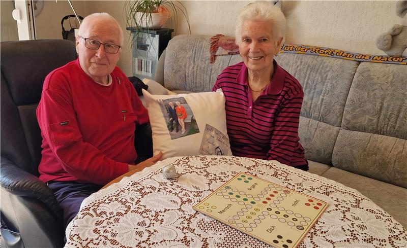 Zwei ältere Personen sitzen an einem runden Tisch mit weißer Spitzen-Tischdecke und einem Spielbrett darauf, im Hintergrund ein graues Sofa und eine Pflanze.