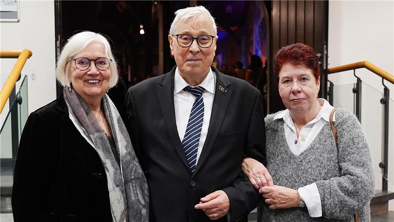 rei Personen stehen nebeneinander in einem Innenraum, der Mann in der Mitte trägt Anzug und Krawatte, die beiden Frauen tragen jeweils einen Pullover und eine Jacke.