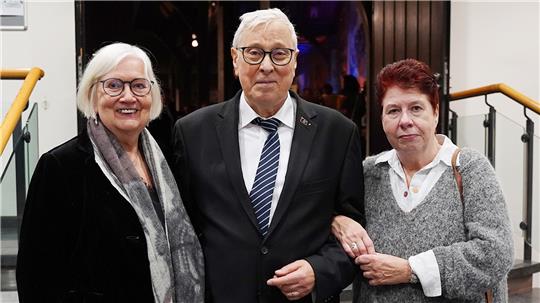 rei Personen stehen nebeneinander in einem Innenraum, der Mann in der Mitte trägt Anzug und Krawatte, die beiden Frauen tragen jeweils einen Pullover und eine Jacke.