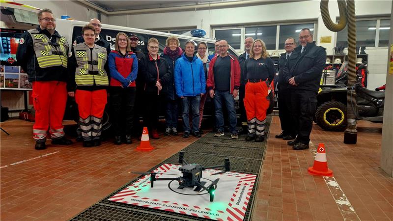 DRK Clausthal-Zellerfeld setzt auf neue Hightech-Drohne Der DRK-Ortsverein präsentiert in seiner Fahrzeughalle den Großspendern die neue Drohne. Das Hightech-Gerät steht im Vordergrund, während sich die Menschen im Halbkreis dahinter aufstellen.