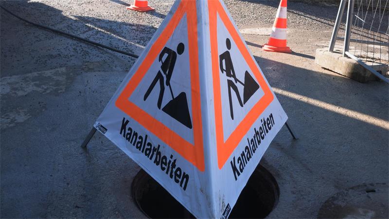 Pyramidenförmiges Warnschild mit orangem Rand und schwarzem Piktogramm eines grabenden Arbeiters sowie dem Schriftzug 'Kanalarbeiten' vor einer offenen Kanalöffnung auf Asphalt.