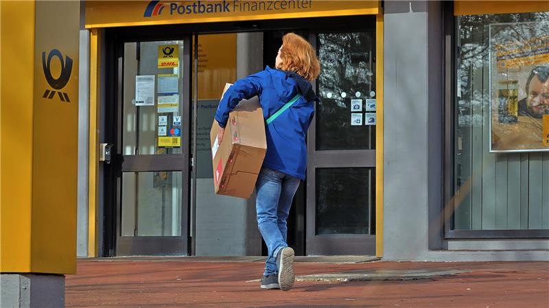 Person mit blauer Jacke und Jeans trägt ein Paket und geht auf einen Eingang mit Postbank-Schriftzug zu