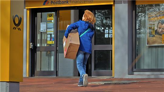 Person mit blauer Jacke und Jeans trägt ein Paket und geht auf einen Eingang mit Postbank-Schriftzug zu