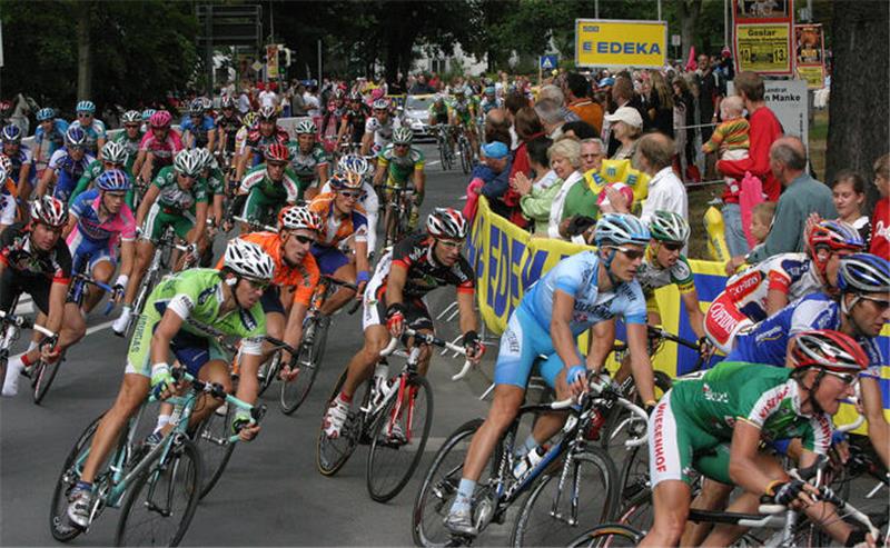 Gruppe von Radrennfahrern in bunter Sportkleidung fährt auf einer Straße, umgeben von Zuschauern hinter Absperrungen mit Werbebannern