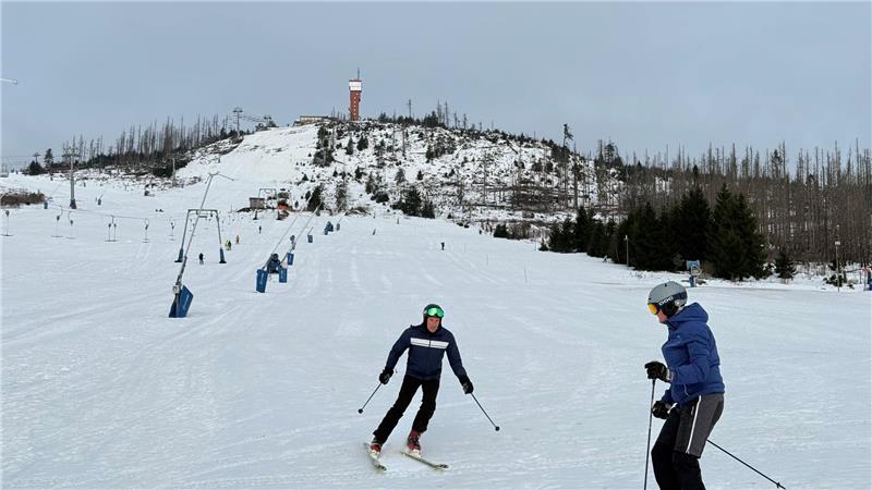 Zwei Skifahrer auf einer schneebedeckten Piste mit Sessellift und bewaldetem Hügel im Hintergrund.