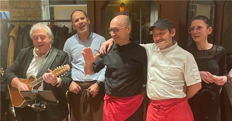 Fünf Männer in Kochkleidung, zwei mit roten Schürzen, einer spielt Mandoline, sie stehen in einem Raum mit Garderobe im Hintergrund.