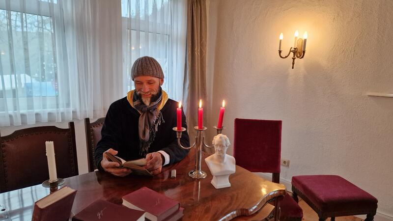 Achim Kapelle sitzt an einem Tisch und liest aus einem von Goethes Werken. Vor ihm auf dem Tisch und hinter ihm an der Wand brennen Kerzen. Auf dem Tisch steht eine Büste von Goethe. 