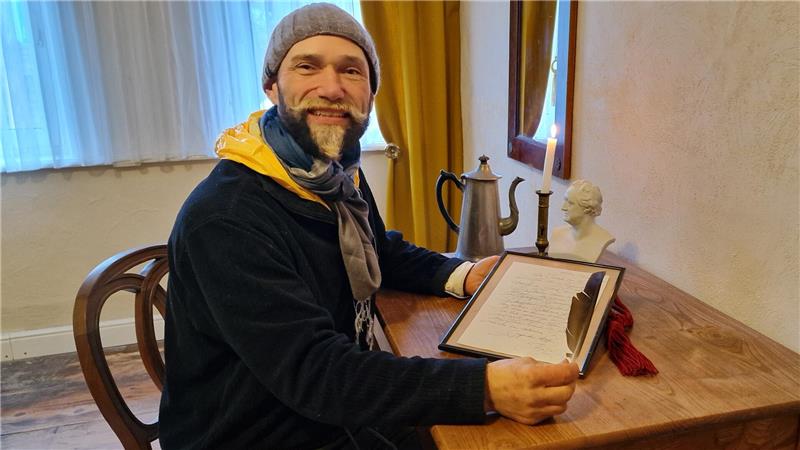 Das Bild zeigt Achim Kapelle, auf einem Stuhl. In der Hand hält er ein Bilderrahmen mit einem Schriftstück und eine Feder. Im Hintergrund brennt eine Kerze und eine Büste von Goethe steht auf dem Tisch. 