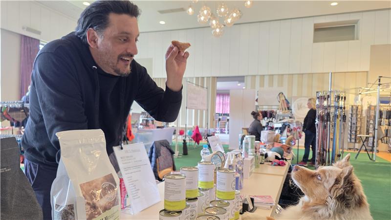 Mann steht an einem Tisch mit Produkten und hält ein kleines Stück in der Hand, Hund sitzt davor und schaut nach oben
