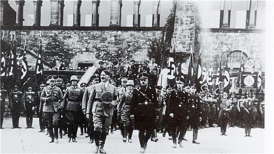 Schwarz-weiß-Foto einer militärischen Parade mit mehreren uniformierten Männern und zahlreichen Hakenkreuzfahnen vor einem steinernen Gebäude.