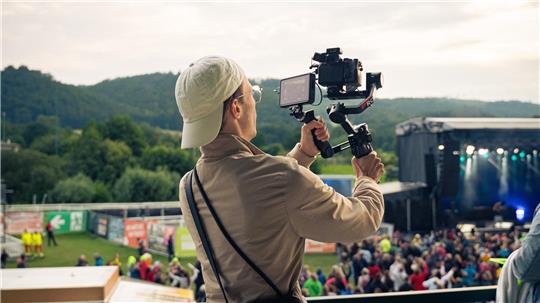 Ein Mann mit umgedrehter Cap auf dem Kopf hält eine Kamera hoch und filmt eine Bühne, vor der Publikum steht.