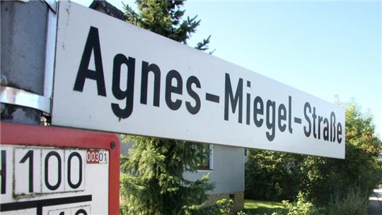 Straßenschild mit der Aufschrift 'Agnes-Miegel-Straße' vor Bäumen und einem Haus im Hintergrund