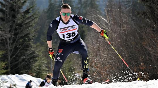 Biathlet in dunklem Anzug mit Startnummer 136 beim Wettkampf auf schneebedeckter Strecke vor Nadelwald.
