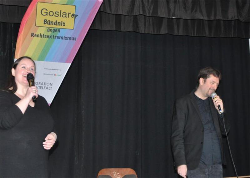 Zwei Personen mit Mikrofonen vor schwarzem Bühnenvorhang, links ein Banner mit Regenbogenfarben und der Aufschrift 'Goslarer Bündnis gegen Rechtsextremismus'