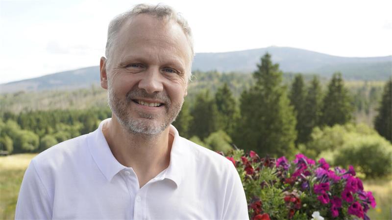 Mann mit kurzem grauem Haar und Bart in weißem Poloshirt vor Landschaft mit Bäumen und Blumen.