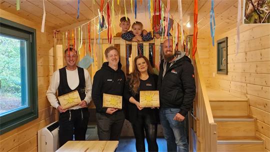 Fünf Erwachsene mit Holzplatten in einem Holzraum, zwei Kinder schauen von einer Galerie herunter.