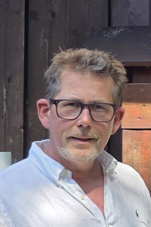 Mann mit kurz geschnittenem Haar trägt ein weißes Hemd und steht vor einer Holzwand mit einem kleinen Fenster.