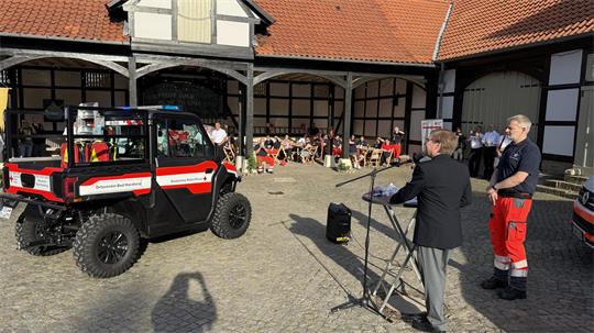 Ein Mann steht am Rednerpult, vor ihm ein geländegängiges Rettungsfahrzeug und im Hintergrund Menschen, die dem Redner zuhören. 