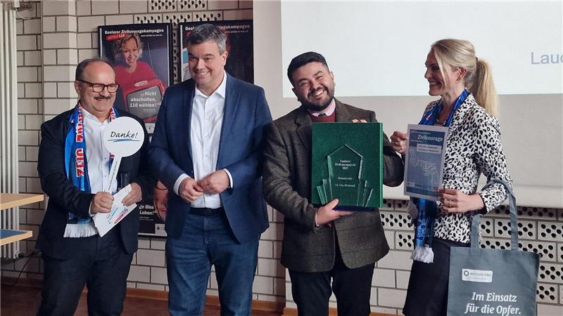 Vier Personen stehen nebeneinander, zwei halten Urkunden, eine trägt einen Schal mit Schrift, alle blicken zur Kamera