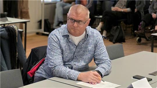 Mann mit grauem Hemd sitzt an einem Tisch in einem Raum mit mehreren Personen im Hintergrund