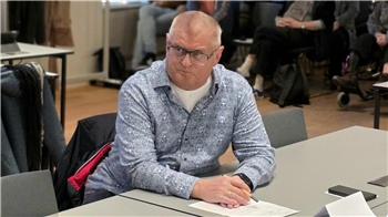 Mann mit grauem Hemd sitzt an einem Tisch in einem Raum mit mehreren Personen im Hintergrund