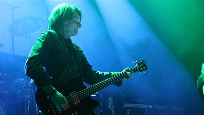 Musiker auf Bühne spielt E-Bass unter grün-blauem Bühnenlicht, im Hintergrund Schlagzeug und Verstärker sichtbar.