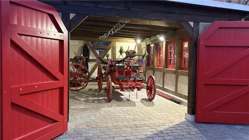 Pferdespritze nimmt ihren Platz im Kleinen Spritzenhaus ein Offene rote Doppeltore führen zu einem Hof mit historischen roten Feuerwehrwagen unter einem Holzdach vor einem Fachwerkhaus.