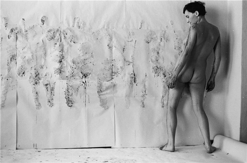 Ein nackter, mit Farbe eingeschmierter Mann dreht sich an einer Papierwand um die eigene Achse und hinterlässt auf dem Papier Farbabdrücke seines Körpers.