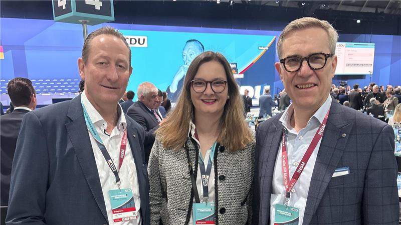 Drei Personen mit Namensschildern auf einer Veranstaltung vor einer großen blauen Leinwand mit Text und einem Logo