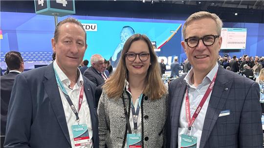 Drei Personen mit Namensschildern auf einer Veranstaltung vor einer großen blauen Leinwand mit Text und einem Logo