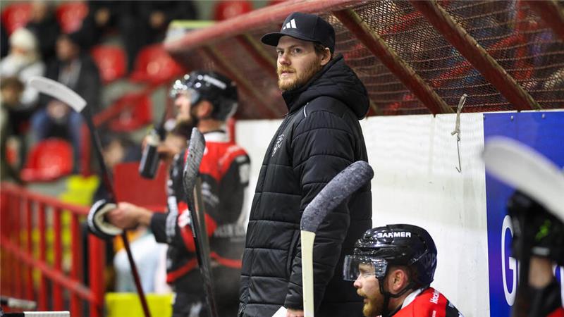 Person in schwarzer Winterjacke und Mütze steht an der Bande neben sitzendem Eishockeyspieler in rotem Trikot mit Helm