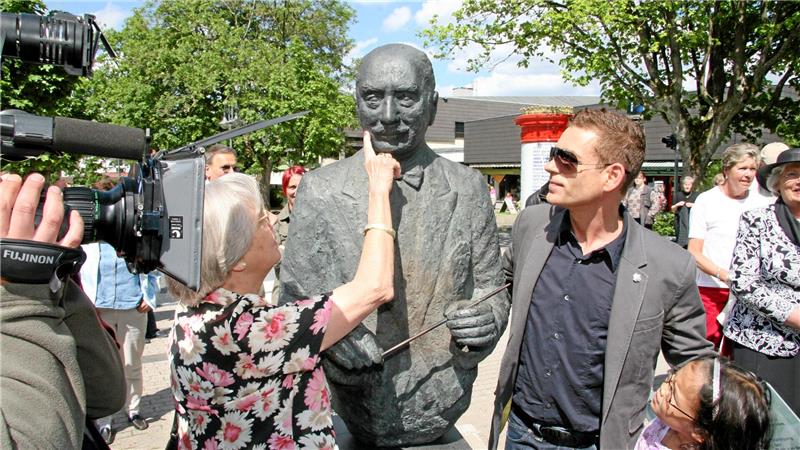 Eine Frau berührt die Brust einer Bronzestatue eines Mannes, umgeben von mehreren Personen und einem Kameramann mit einer großen Kamera im Freien.