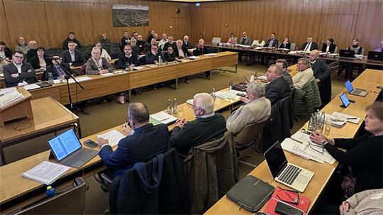 Mehrere Menschen, die in einem Saal an langen Tischen sitzen. 
