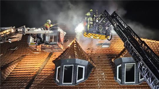 Feuerwehrleute auf zwei Drehleitern löschen nachts ein Feuer auf einem Ziegeldach mit Gaubenfenstern.