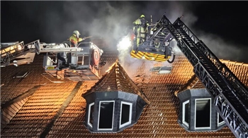 Feuerwehrleute auf zwei Drehleitern löschen nachts ein Feuer auf einem Ziegeldach mit Gaubenfenstern.