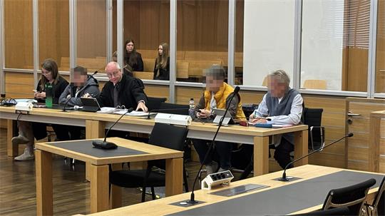 Mehrere Personen sitzen an einem langen Tisch in einem Raum mit Holzvertäfelung, einige Gesichter sind unscharf.
