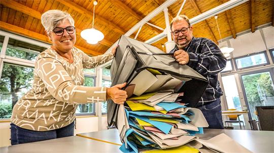 Das Bild zeigt eine Frau und einen Mann, die aus einer Wahlurne die Stimmzettel auf einem Tisch ausschütten.