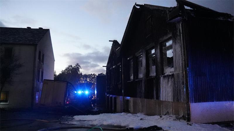Ausgebrannte Industriehalle nach einem Großbrand bei Dunkelheit, mit Einsatzfahrzeugen und Blaulicht im Hintergrund.