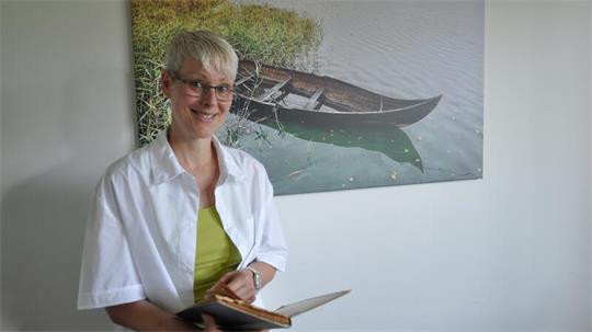 Person mit kurzem blondem Haar, weißem Hemd und grünem Oberteil hält ein aufgeschlagenes Buch vor einem Bild mit einem Boot auf ruhigem Wasser.